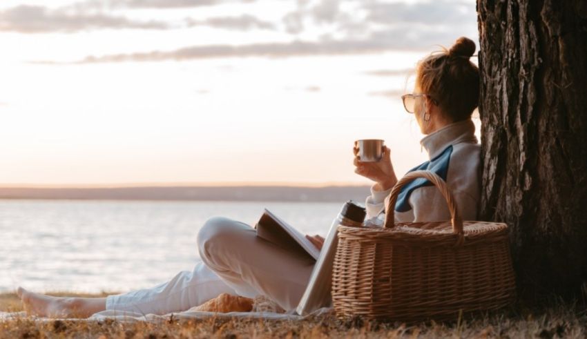 A woman is sitting under a tree reading a book and drinking coffee.