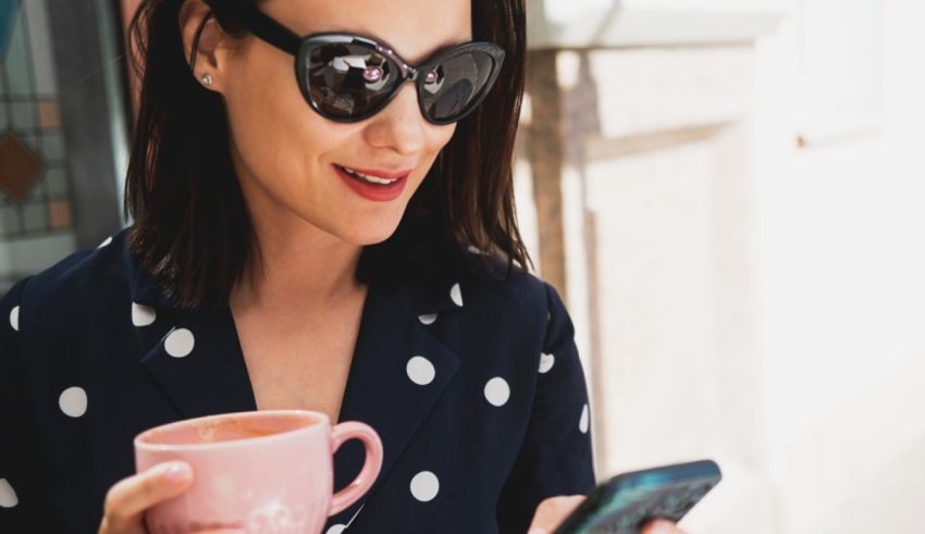 A woman in sunglasses holding a cup of coffee and a cell phone.