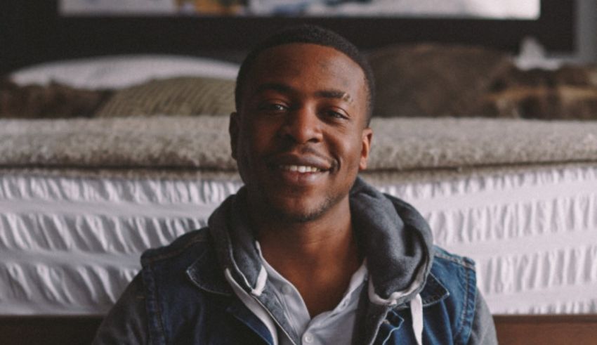 A man in a denim jacket sitting on a bed.