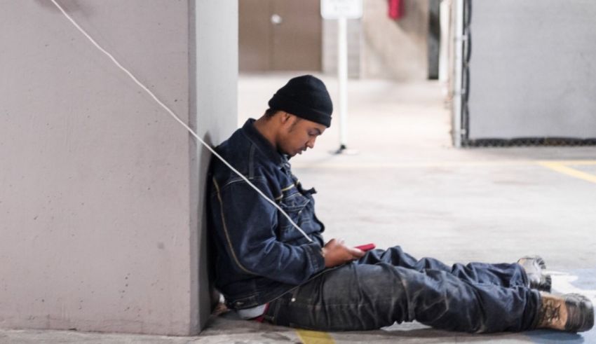 A homeless man sitting on the ground with a cell phone.