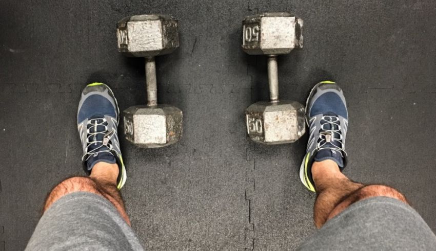 A person's legs standing on a gym floor with two dumbbells.