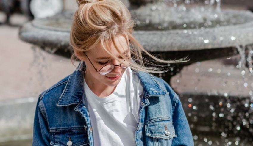 A woman wearing glasses and a denim jacket in front of a fountain.