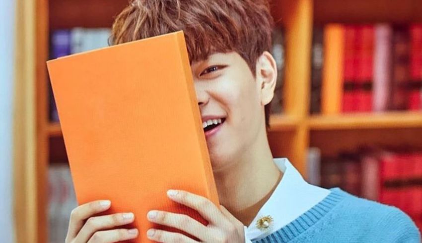 A young man holding an orange book in front of a bookshelf.