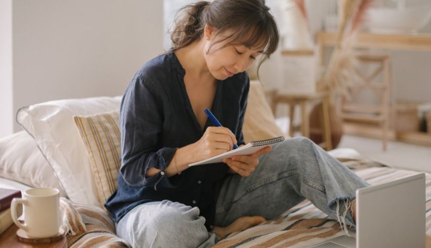 A woman writing on a laptop while sitting on a bed.