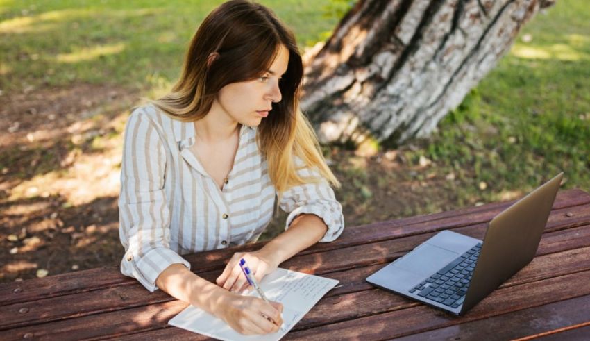 A young woman sitting at a picnic table with a laptop and a notebook.