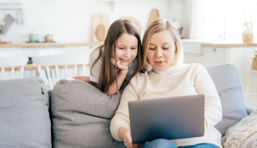 A woman and her daughter are sitting on a couch looking at a laptop.