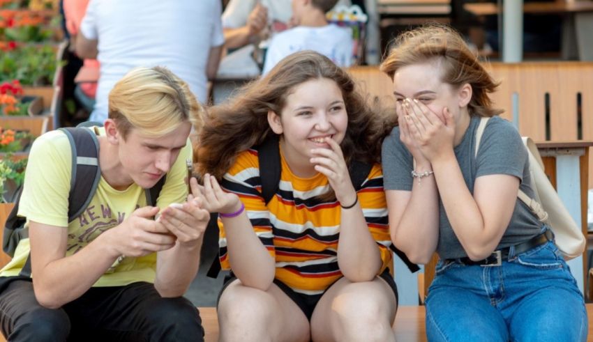 Three young people sitting on a bench looking at their cell phones.