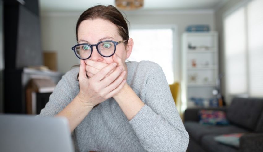 A woman with glasses covering her mouth while using a laptop.