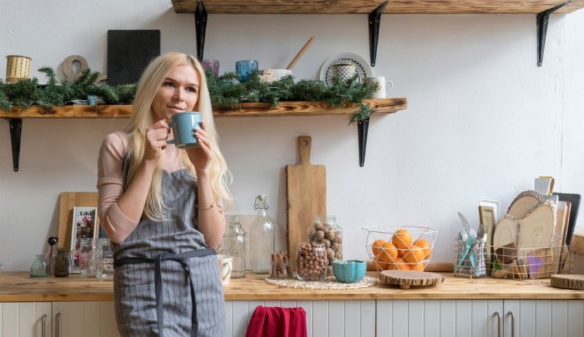 A blonde woman in an apron is holding a coffee mug in her kitchen.