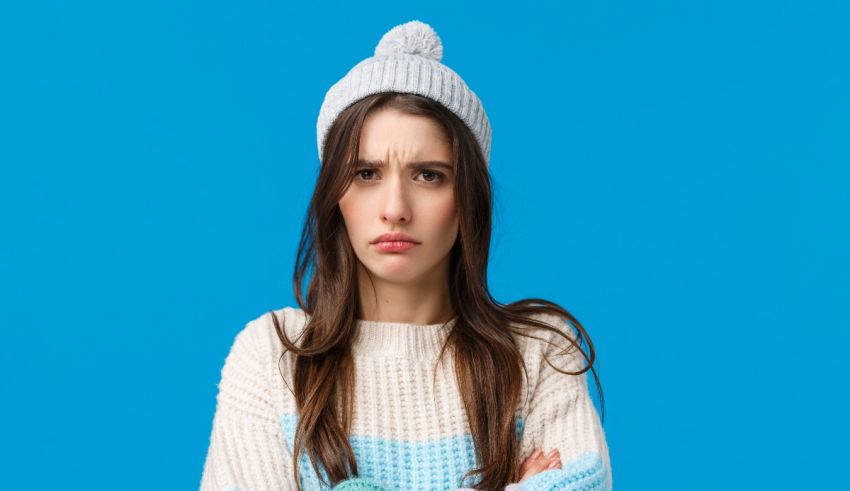 A young woman wearing a sweater and a beanie on a blue background.