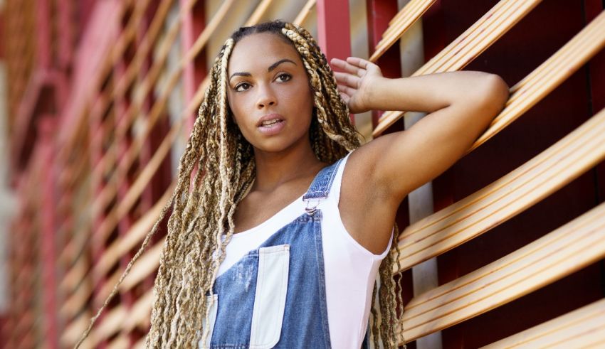 A black woman in overalls leaning against a wooden wall.