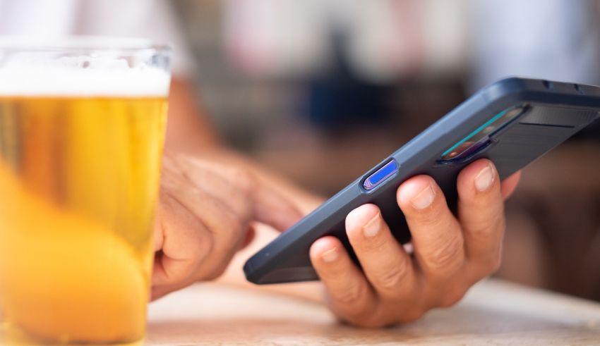 A person holding a cell phone next to a glass of beer.