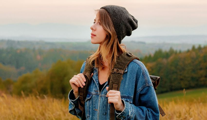 A woman in a denim jacket standing in a field with a backpack.