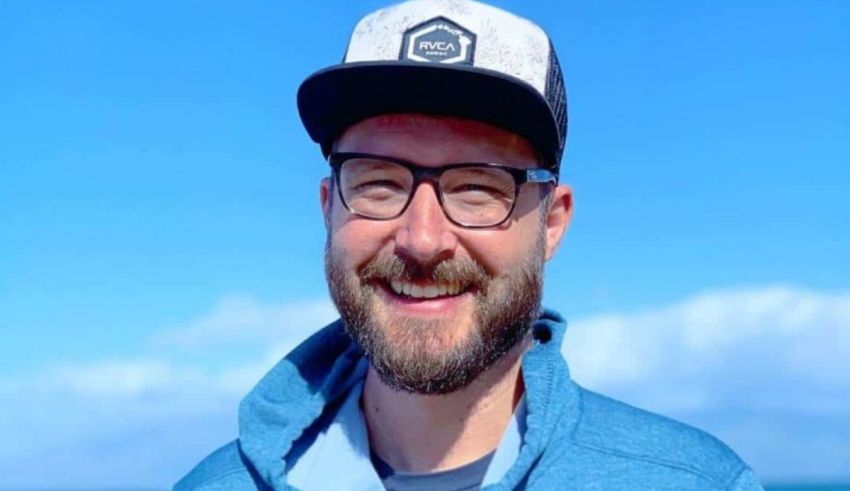 A man with glasses and a hat smiling in front of the ocean.