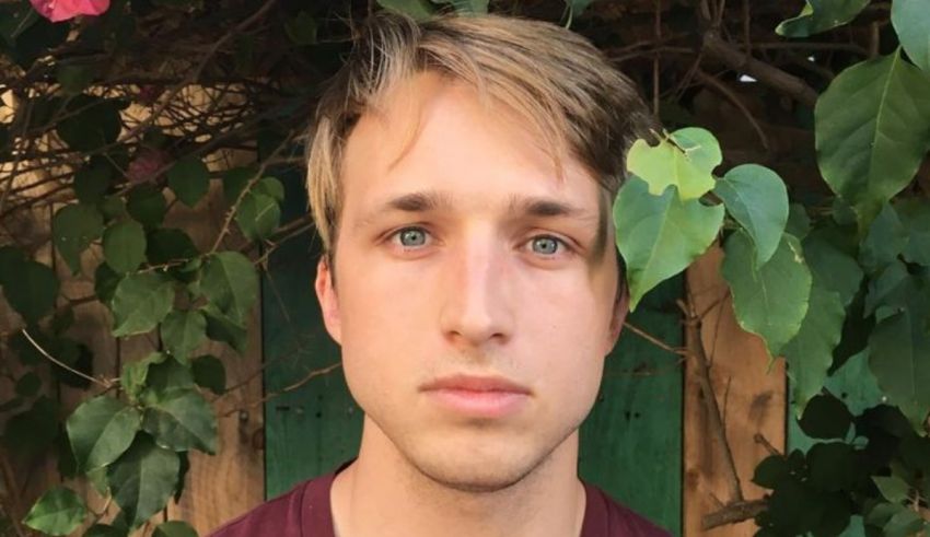 A young man in a maroon shirt is standing in front of some plants.