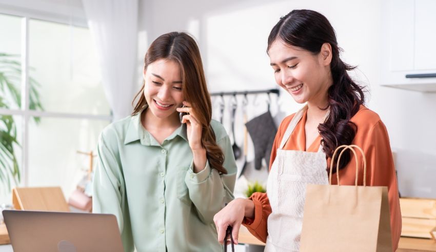 Two asian women talking on the phone while holding shopping bags.