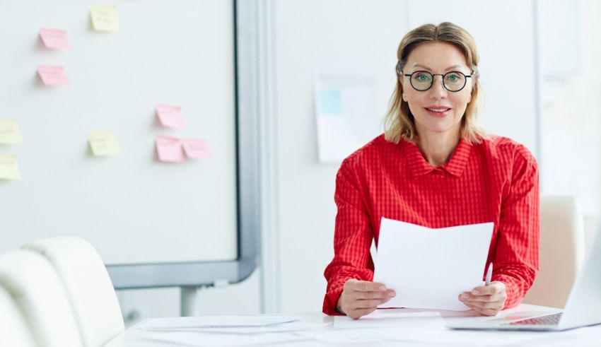 A woman wearing glasses is sitting at a desk and holding a piece of paper.