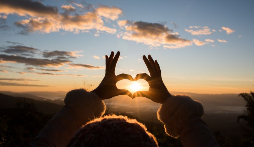 A woman's hands make a heart shape at sunset.