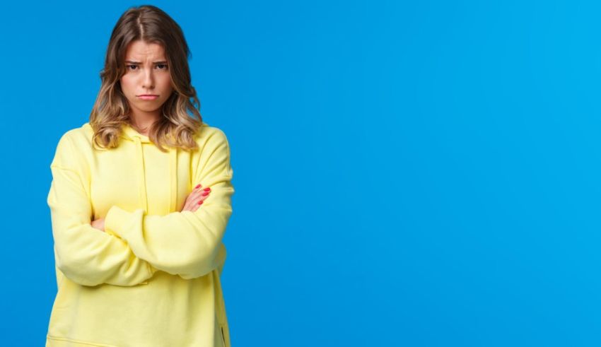 A young woman in a yellow sweatshirt standing with her arms crossed on a blue background.