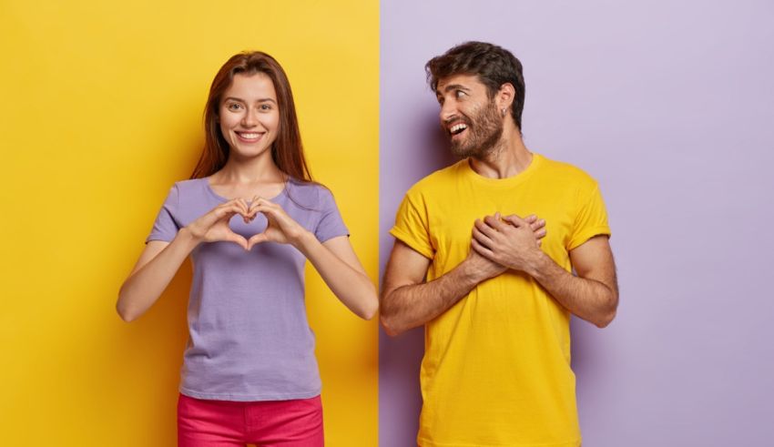 Young man and woman making a heart shape on a yellow and purple background.