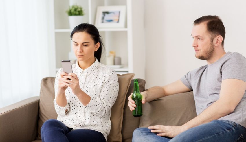 A man and woman sitting on a couch with a bottle of beer and a cell phone.