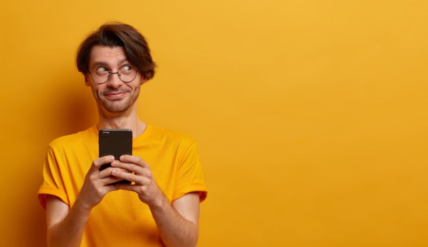 A man with glasses is holding a smart phone against an orange background.