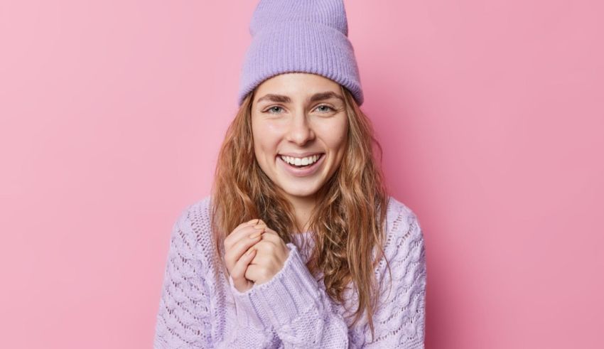 A young woman wearing a purple beanie and sweater on a pink background.