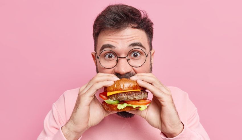 A man with glasses is eating a hamburger on pink background.