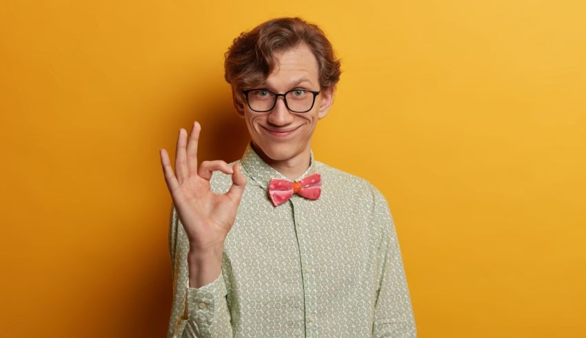 A young man with glasses and a bow tie showing the ok sign on a yellow background.