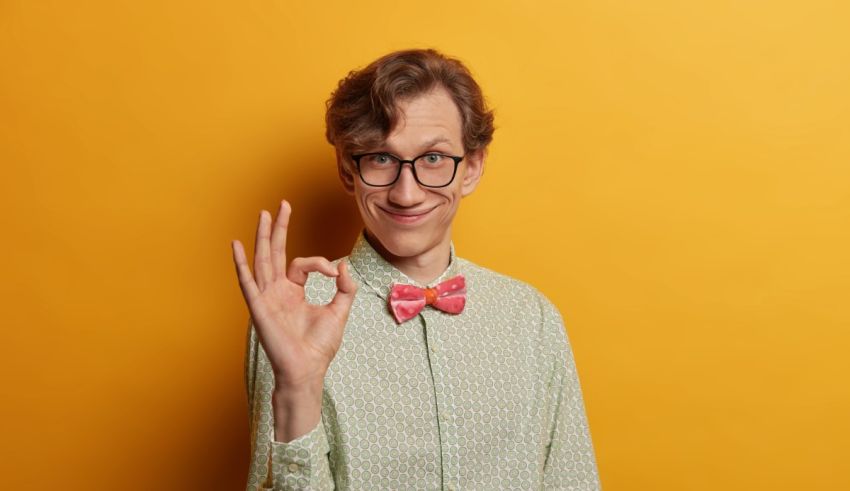 A young man with glasses and a bow tie showing the ok sign on a yellow background.