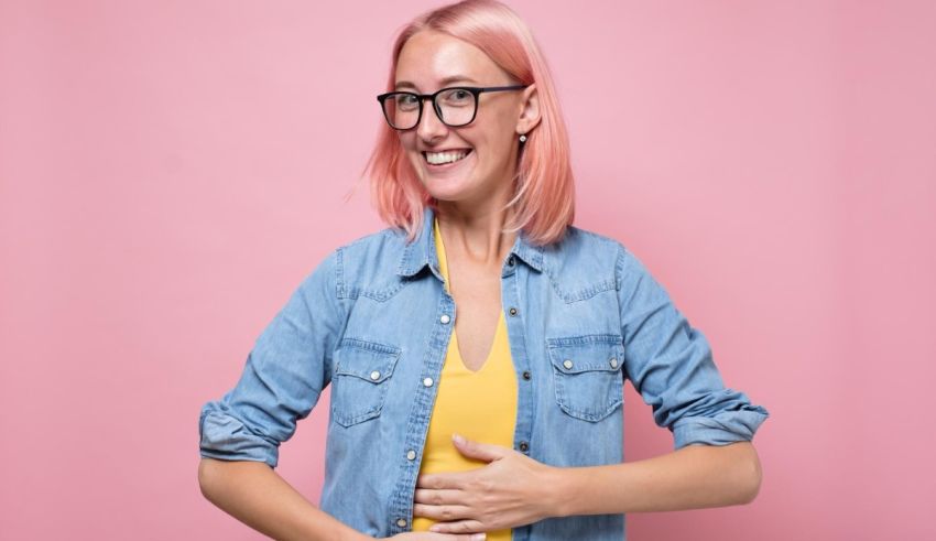 A woman with pink hair and glasses holding her stomach against a pink background.
