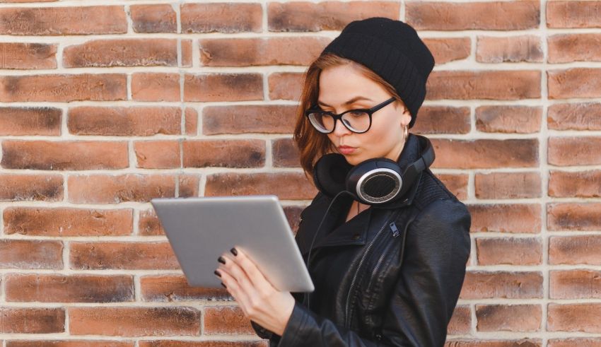 A young woman wearing glasses and a beanie leaning against a brick wall using a tablet computer.