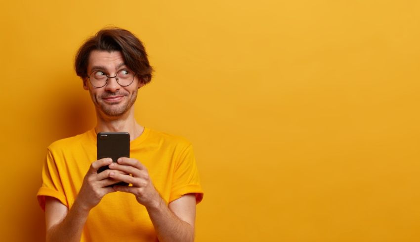A man with glasses is holding a smart phone against a yellow background.