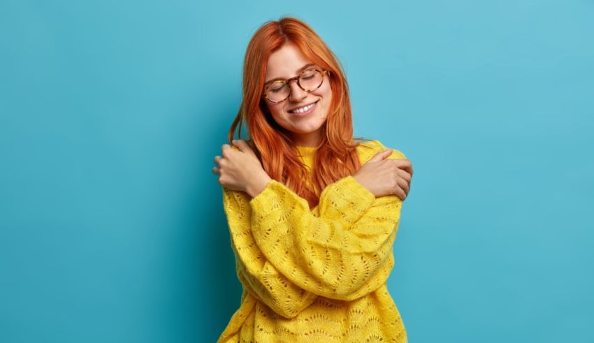 A young woman with red hair and glasses is posing against a blue background.