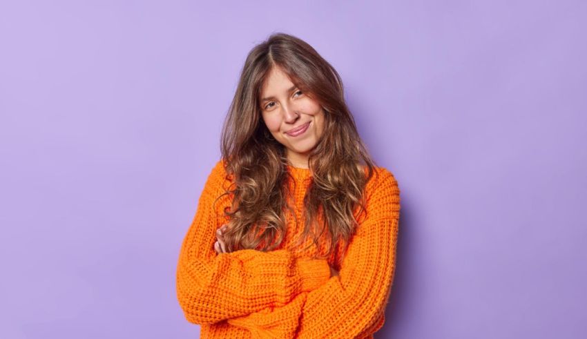 A young woman in an orange sweater is posing against a purple background.