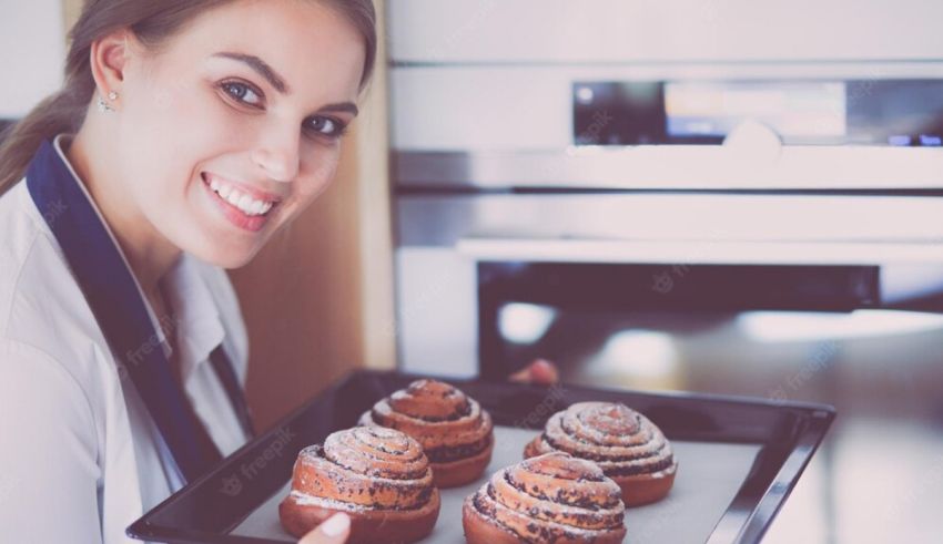 A woman is holding a tray of cinnamon buns in front of an oven.