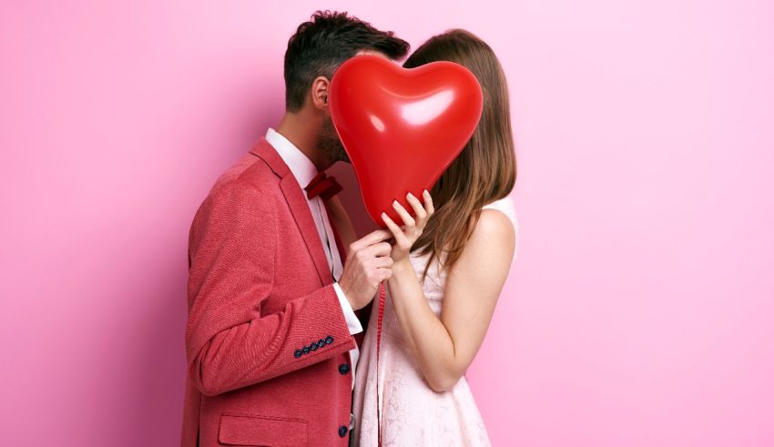 A man and woman holding a red heart balloon against a pink background.