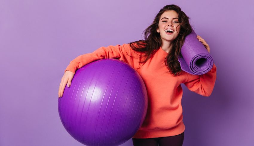 A young woman is holding a purple exercise ball on a purple background.