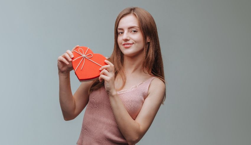 A young woman holding a heart shaped box on a gray background.