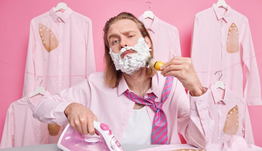 A man with a beard is ironing clothes on a pink background.