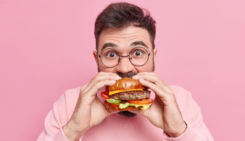 A man with glasses is eating a hamburger on a pink background.