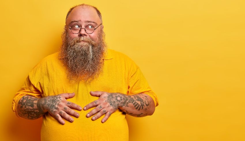 A bearded man with tattoos and glasses standing on a yellow background.