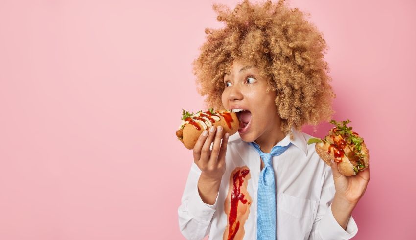 A young woman eating a hot dog with ketchup on a pink background.