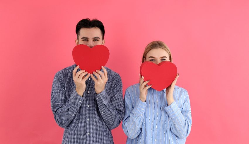 Young man and woman holding red heart shaped paper over pink background.