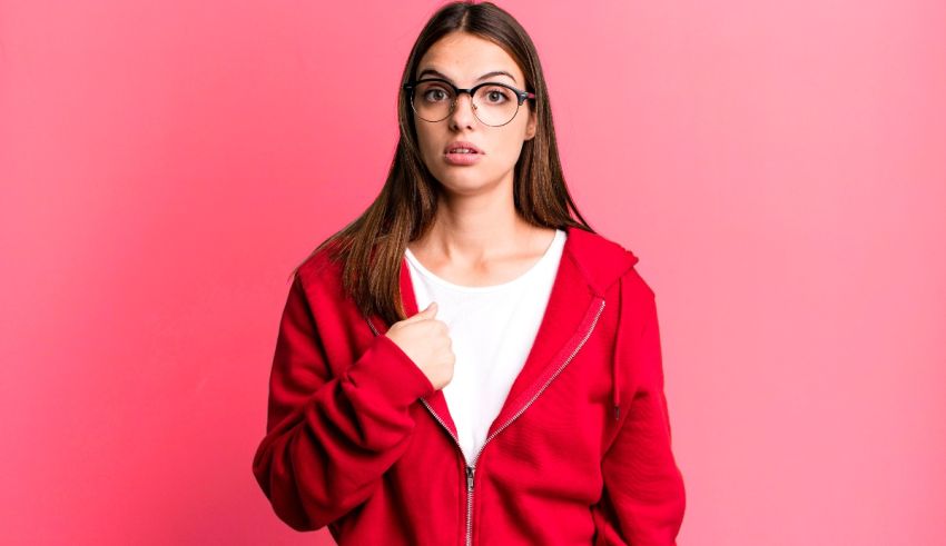 A woman wearing glasses and a red hoodie is standing on a pink background.