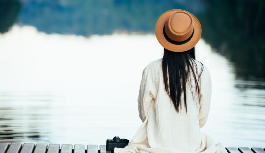A woman wearing a hat sits on a dock next to a lake.