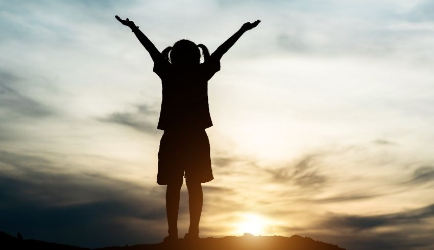 A silhouette of a girl standing on top of a hill with her arms raised.