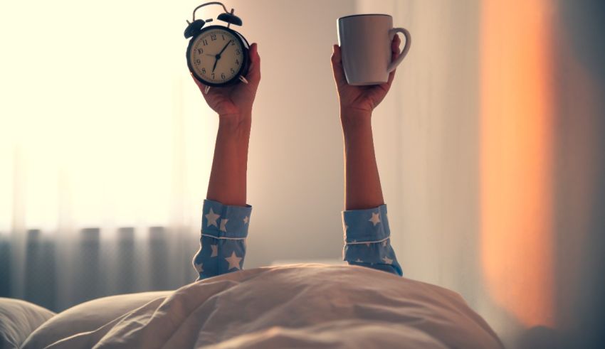 A woman laying in bed with an alarm clock and a cup of coffee.