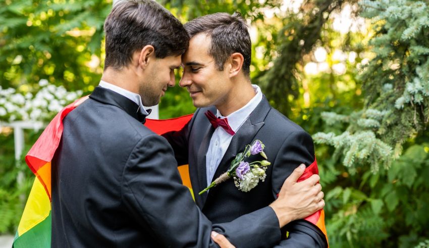 Two men in tuxedos hugging each other with a rainbow flag.