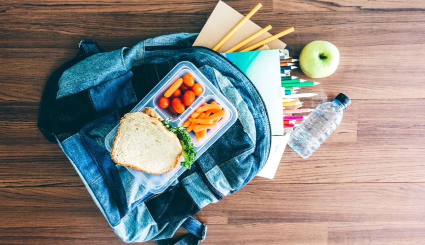 A backpack with a sandwich, apples and pencils on a wooden floor.